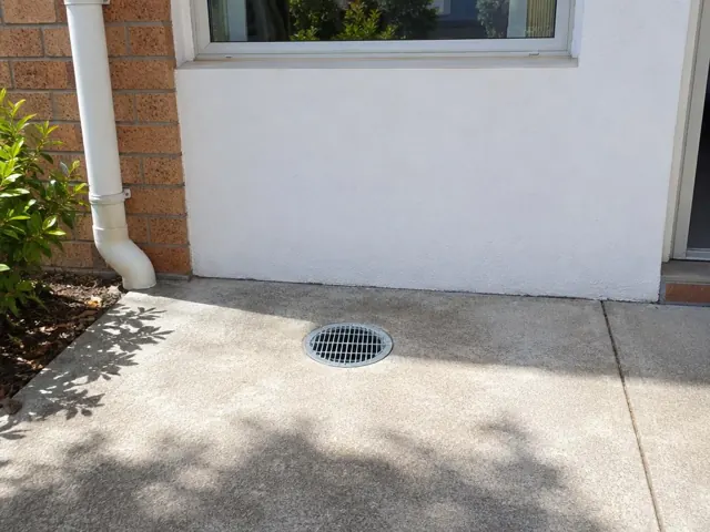 Outdoor drain grate set in dry concrete paving beside a brick wall and house exterior in Abbotsbury