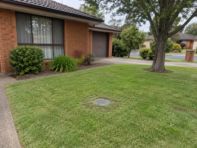 Suburban front lawn with a flush sewer access cap beside a brick house in Prairiewood